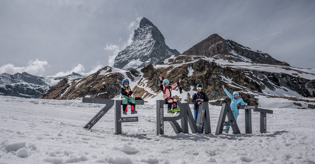 family skkinging in zermatt switzerland sitting on the large Zermatt letters infront of the Matterhorn