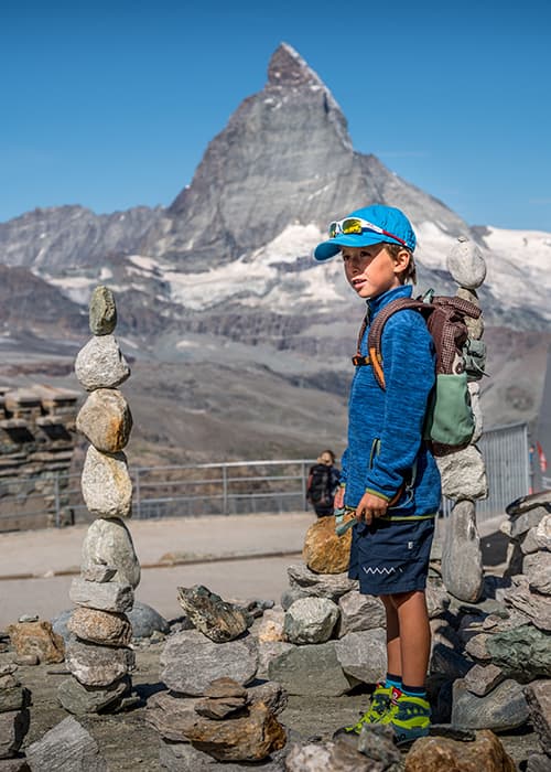 a boy in a blue jacket standing at the gornergrat infront of the matterhorn