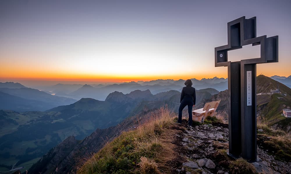 a woman standing during sunrise at the top of the mountain Rothorn next to a cross