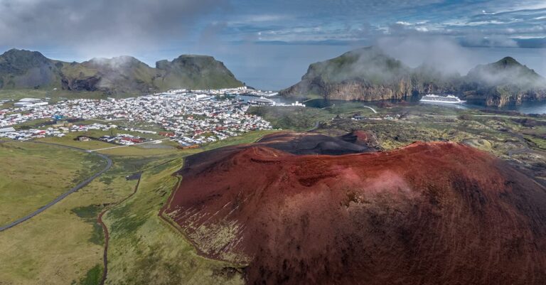 areas view of a dormant volcano with a village in the background and Rocky Mountains in Icleand